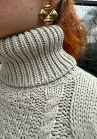 Gros plan d'une personne portant un pull à col roulé épais en maille torsadée gris clair et de grandes boucles d'oreilles carrées texturées en or.