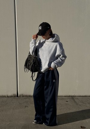 Sudadera gris con un bolsillo frontal, combinada con pantalones de chándal navy con rayas blancas. Gorra negra y gafas de sol, sosteniendo un bolso negro.