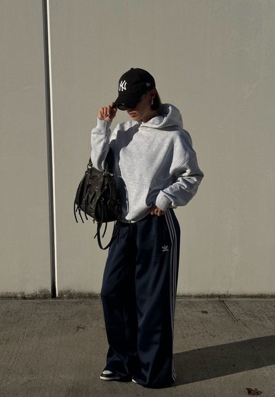 Sudadera gris con un bolsillo frontal, combinada con pantalones de chándal navy con rayas blancas. Gorra negra y gafas de sol, sosteniendo un bolso negro.