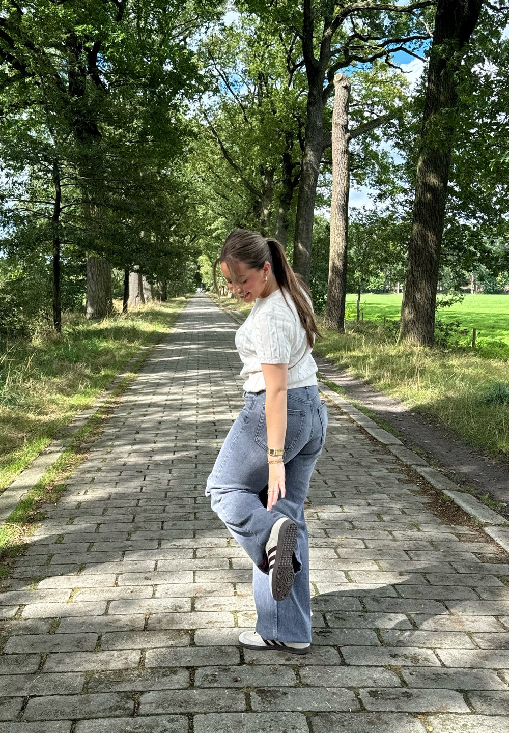 Conjunto casual com um suéter creme de malha trançada e jeans de cintura alta azuis. Cenário numa senda rodeada de árvores com pavimento de pedras.