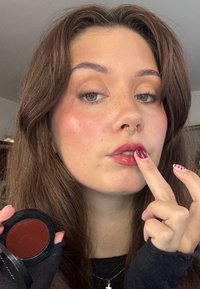 Okrągłe czarne opakowanie zawierające głęboko czerwony krem. Osoba aplikuje produkt na usta palcem, ukazując błyszczące wykończenie i zadbane paznokcie.