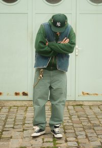 Gilet en denim par-dessus un sweat à capuche vert, pantalon vert ample, baskets noires et blanches. Porte-clés attaché au pantalon. En arrière-plan, une porte vert menthe.