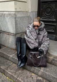 Szary płaszcz z futrzanym kapturem, czarne skórzane kozaki za kolano, ciemna torebka i czarne rękawiczki, siedząca na kamiennych schodach na tle o fakturowanej powierzchni.