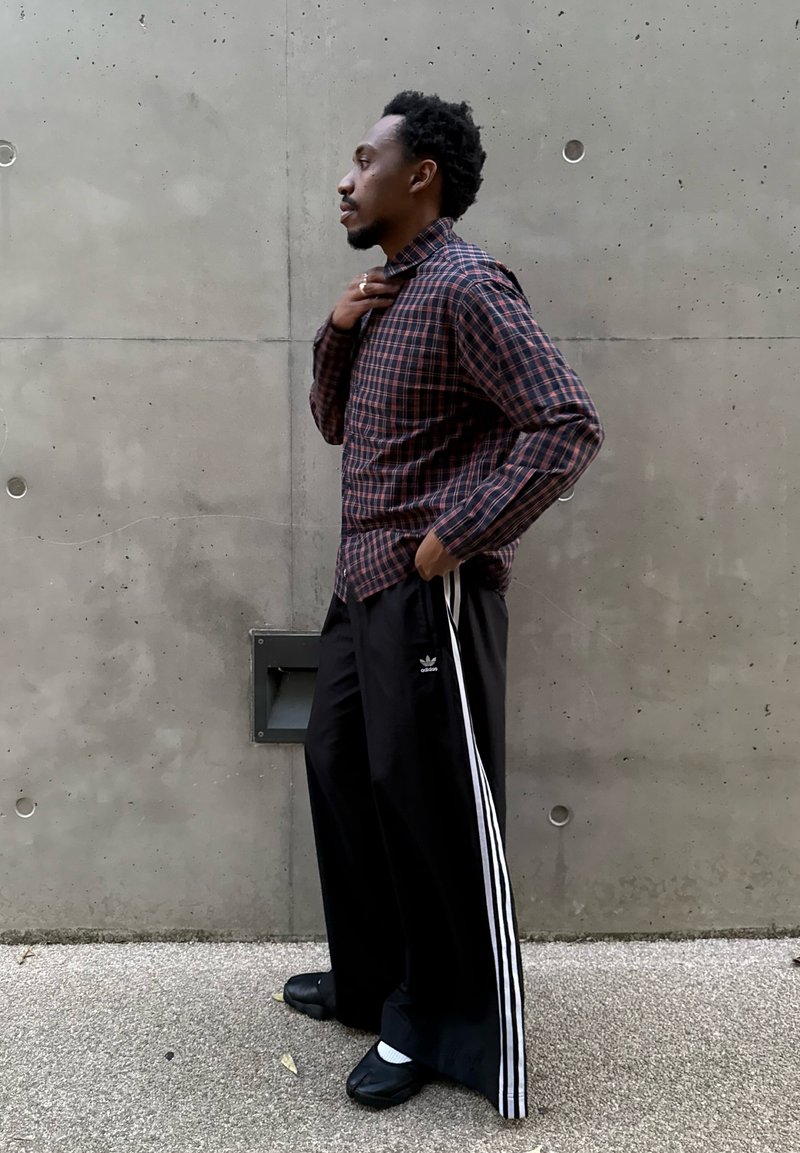 Homme debout de côté, portant une chemise à carreaux, un pantalon de survêtement Adidas noir avec des rayures blanches, et des baskets noires, devant un mur en béton.