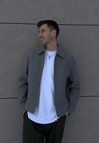 Veste grise zippée en tissu texturé, portée sur une chemise blanche. Détails remarquables : col pointu et poches latérales.