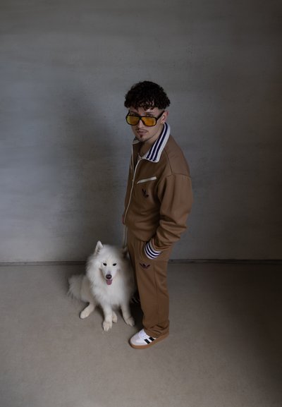 Chándal marrón con cuello a rayas blancas y moradas, combinado con zapatillas blancas con rayas negras. Un perro blanco y esponjoso se sienta al lado.