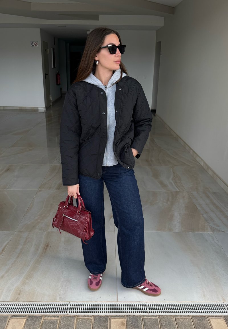 Femme portant une veste matelassée noire, un sweat à capuche gris, un jean large foncé, des baskets roses, des lunettes de soleil noires, tenant un sac à main rouge, debout dans un couloir.