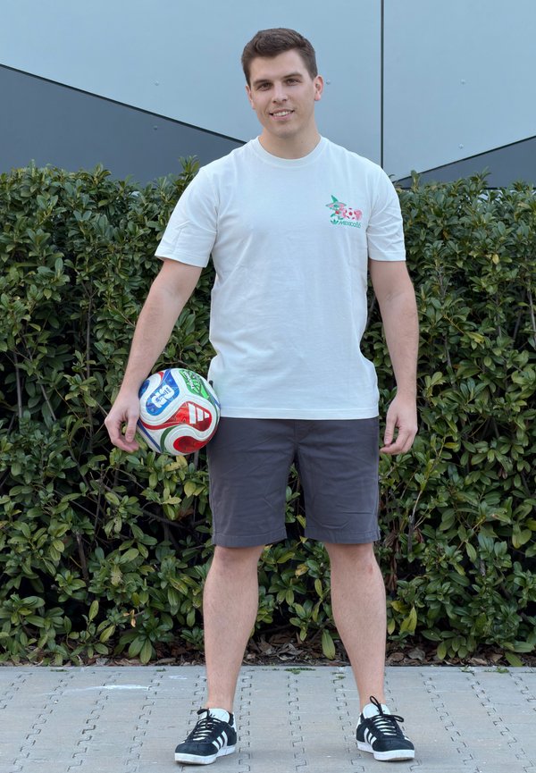 Joven con camiseta blanca y pantalones cortos oscuros sosteniendo un balón de fútbol a su lado, de pie en un sendero pavimentado con arbustos verdes detrás de él.