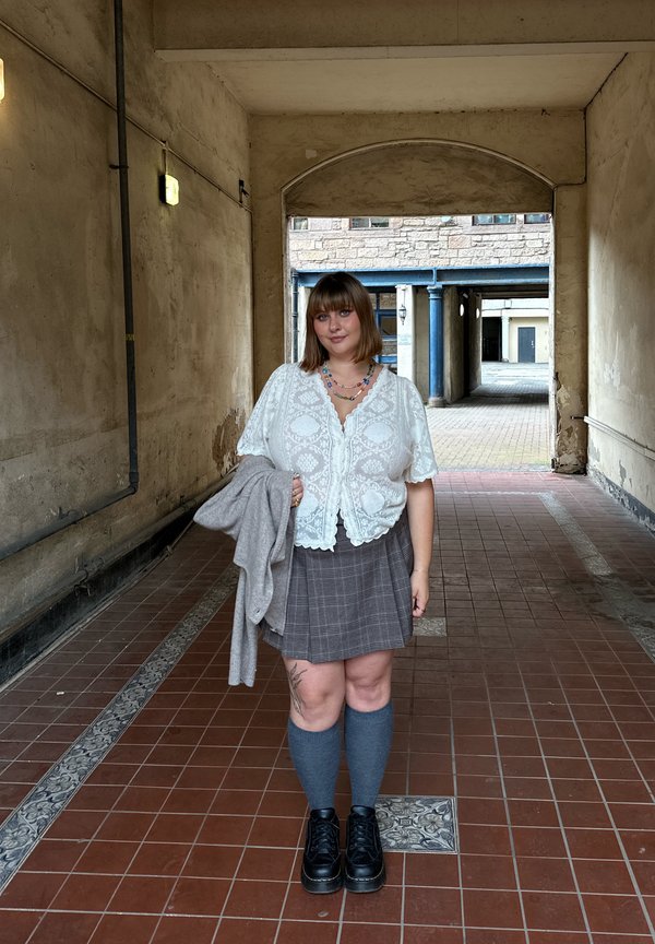 Jeune femme debout dans une arche carrelée portant un top en dentelle blanc, une jupe grise à carreaux, des chaussettes montantes, des chaussures noires et tenant une veste grise.