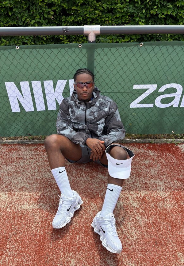 Mann in Sportbekleidung und Sonnenbrille sitzt auf einem roten Tennisplatz vor einem grünen Zaun mit Nike-Logo und hält ein weißes Nike-Visier.