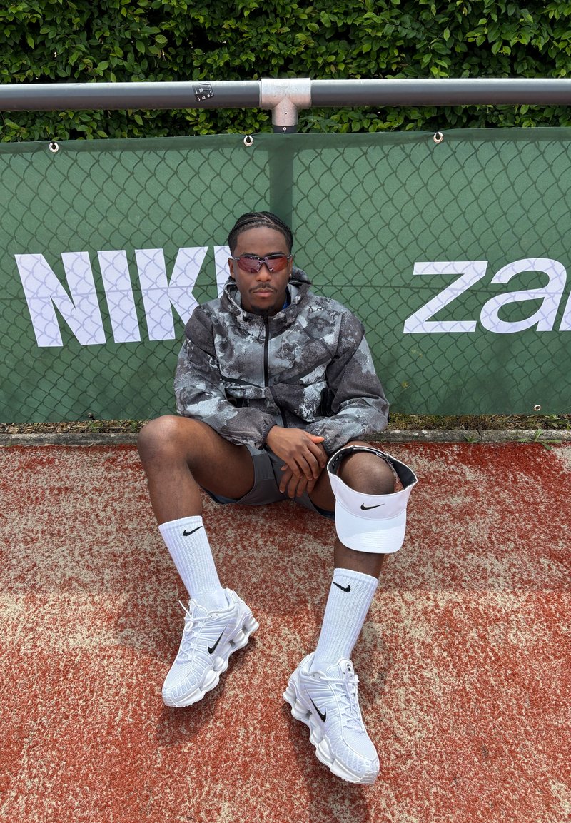 Homme en tenue de sport et lunettes de soleil assis sur un court de tennis rouge devant une haie verte avec le logo Nike, tenant une visière blanche Nike.