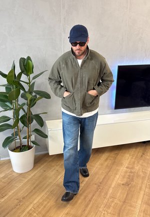 Hombre con gafas de sol oscuras, gorra azul marino, chaqueta verde y vaqueros azules, caminando en el interior cerca de un televisor y una maceta grande sobre suelo de madera.