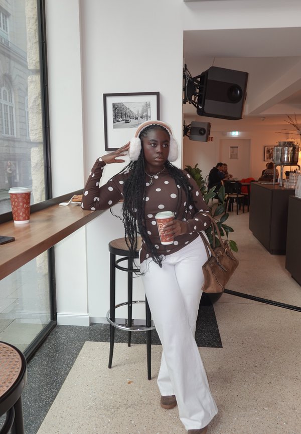 Brown polka dot long-sleeve top, white flared trousers, fuzzy pink earmuffs, holding a coffee cup, standing beside a wooden bar in a café.