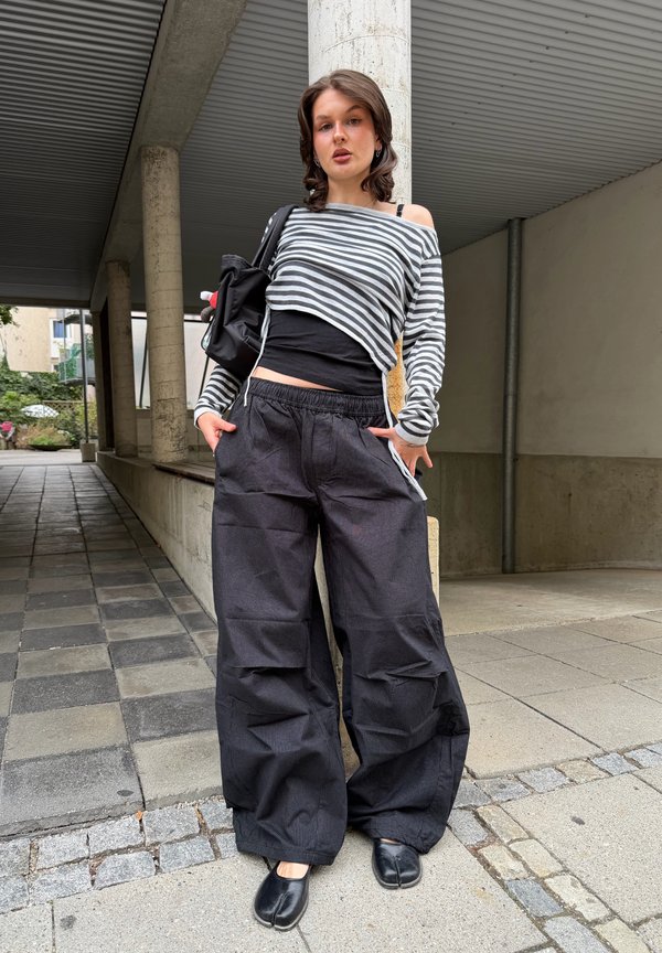 Joven mujer que lleva una camiseta de manga larga a rayas, pantalones anchos negros, zapatos negros y lleva una mochila negra, de pie al aire libre junto a una columna.