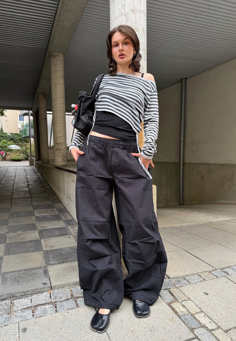 Joven mujer que lleva una camiseta de manga larga a rayas, pantalones anchos negros, zapatos negros y lleva una mochila negra, de pie al aire libre junto a una columna.