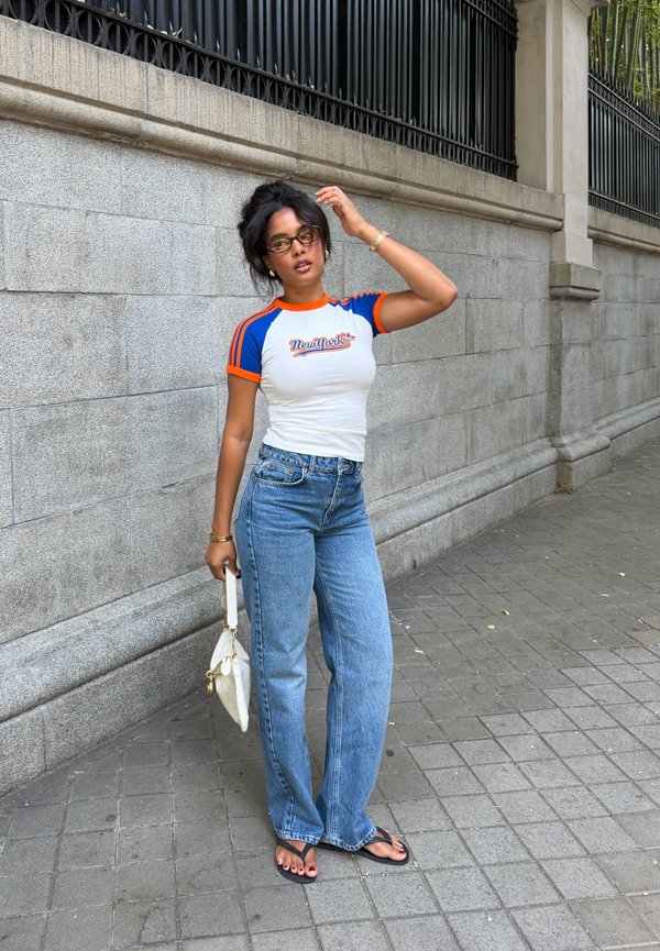 Femme portant des lunettes, un t-shirt New York blanc et bleu, un jean bleu et des sandales, tenant un sac à main blanc, debout près d'un mur en pierre avec une clôture en fer noir.