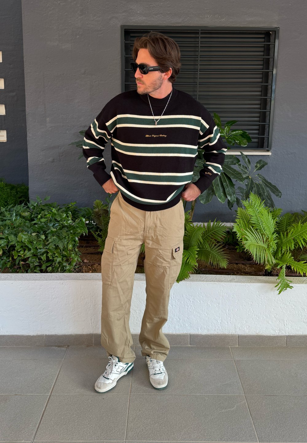 Sweater de malha risca preta e verde com decote redondo, calças cargo bege e ténis brancos, de pé à frente de plantas verdes.