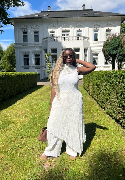 Vestido blanco texturizado con un dobladillo asimétrico combinado con pantalones blancos. Los accesorios incluyen un bolso marrón y gafas de sol oscuras. Fondo de paisaje verde.