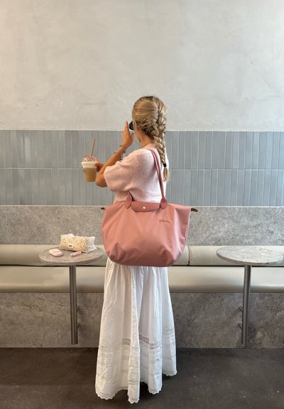 Mujer con trenzas, vestida con un suéter rosa y una falda blanca, sosteniendo un café helado y un teléfono, de pie en un café con un fondo de pared de azulejos grises.