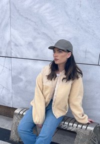Fiatal nő bézs polárdzsekiben, kék farmerben és szürke sapkában, egy fém padon ülve egy szürke kőfal előtt, oldalra nézve.