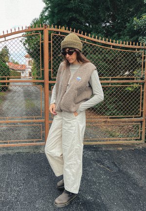 Chaleco de forro polar en marrón claro, camiseta de manga larga gris, pantalones de pierna ancha en color crema, botas marrones y gorro oliva cerca de una puerta de metal.