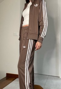 Rudas sportinis kostiumas su baltais dryžiais, sudarytas iš užtraukiamos striukės ir kelnių su elastine juosta. Pagamintas iš lygios medžiagos su logotipo detalėmis.