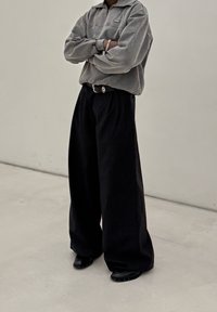 Grau melierter Fleecepullover mit halbem Reißverschluss, schwarze, weit geschnittene Hose mit Gürtel in der Taille und schwarze Slip-On-Schuhe mit Nietenverzierung.