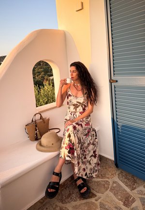 Un vestido maxi de patrón floral en crema con acentos rojos, un bolso de mimbre, un sombrero de sol beige y sandalias de plataforma negras gruesas sobre una superficie de piedra.