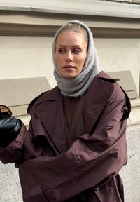 Oversized bruine trenchcoat met brede mouwen, een gestructureerde bruine trui daaronder en een lichtgrijze sjaal. Elegante zwarte handschoenen maken de look compleet.