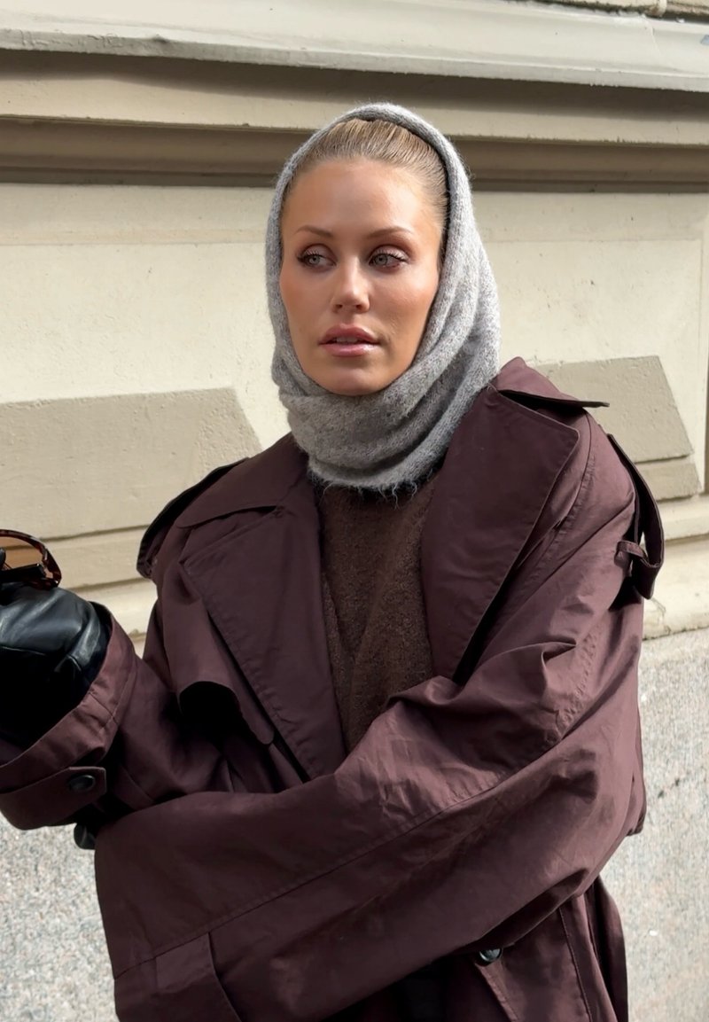 Manteau trench oversized marron avec des manches larges, pull texturé marron en dessous et une écharpe grise claire. Des gants noirs élégants complètent le look.
