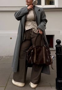 Oversized grijze jas, beige top met strikdetail, bruine wijde broekspijpen, crèmekleurige schoenen en een bruine suède tas met ritsen. Gladde textuur en neutrale kleuren.