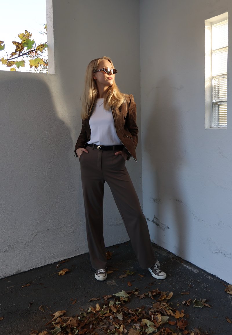 Brun ruskindsjakke over en hvid t-shirt, grå højttaljede bukser og beige sneakers. Stående mod en tekstureret væg, med sollys der skaber skygger.