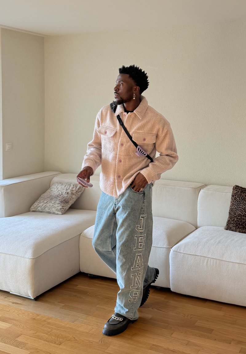 Mann mit einer beige Jacke und Jeans, auf denen "JEANS" auf dem Bein gedruckt ist, steht in einem Wohnzimmer in der Nähe eines weißen Sofas mit Tiermuster-Kissen.