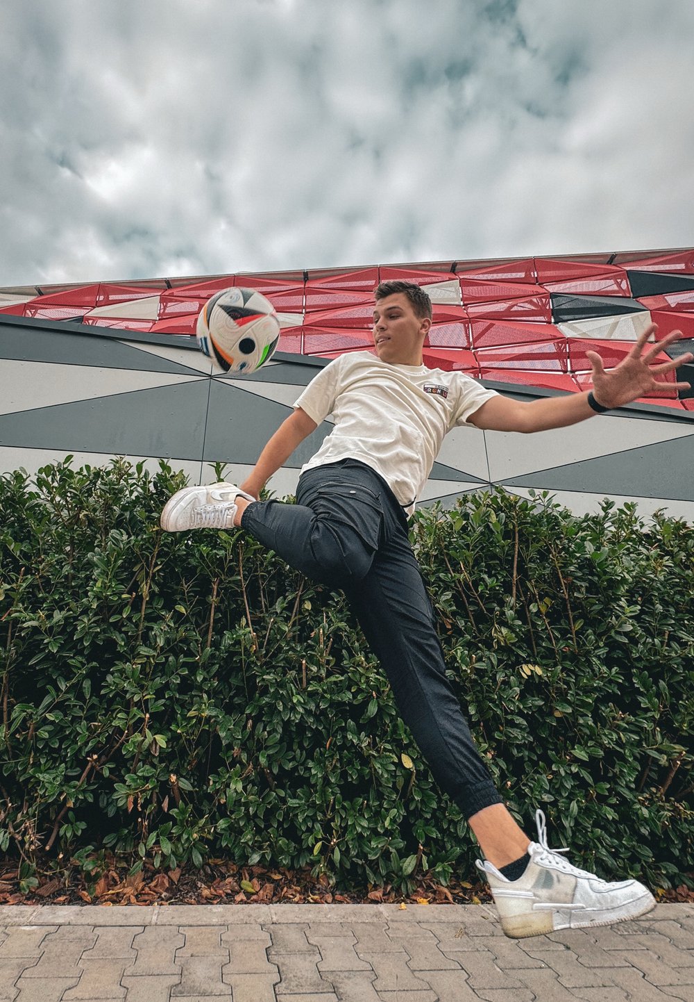 Ένα νέο άτομο με ανοιχτόχρωμο πουκάμισο και μαύρο παντελόνι κλωτσάει μια μαύρη και άσπρη μπάλα ποδοσφαίρου, ανάμεσα σε πράσινα φυτά και έ να κτισμένο φόντο με σχέδια.