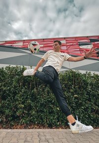 Ένα νέο άτομο με ανοιχτόχρωμο πουκάμισο και μαύρο παντελόνι κλωτσάει μια μαύρη και άσπρη μπάλα ποδοσφαίρου, ανάμεσα σε πράσινα φυτά και ένα κτισμένο φόντο με σχέδια.