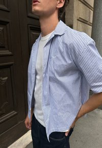 Mężczyzna w niebiesko-białą w paski koszulę z krótkim rękawem, noszącą pod spodem białą koszulkę, stoi z jedną ręką w kieszeni ciemnoniebieskich dżinsów.