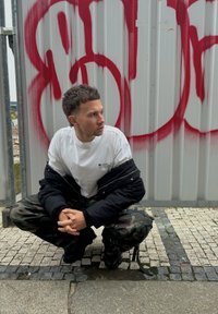 Giubbotto imbottito nero su una maglietta bianca, pantaloni cargo mimetici e sneakers nere. Posizione accovacciata, con graffiti sullo sfondo.