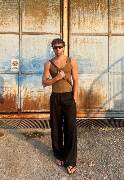 Top sin mangas marrón, pantalones sueltos negros y chanclas. El fondo presenta puertas de metal oxidadas en azul y marrón con un cartel. Sombra en el suelo.