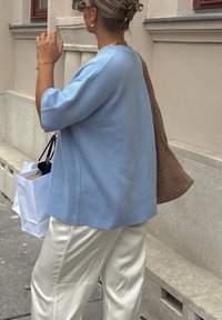 Maglione azzurro chiaro a maglia con maniche corte, abbinato a pantaloni di raso bianchi. Porta borse da shopping bianche e una borsa a tracolla in tessuto intrecciato marrone.
