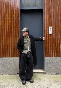 Cappotto lungo nero, camicia verde oliva, T-shirt bianca, pantaloni larghi neri, cappellino grigio, scarpe chunky nere; in piedi accanto a un muro in piastrelle marroni texturizzate.