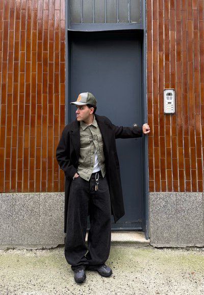 Abrigo largo negro, camisa verde oliva, camiseta blanca, pantalones holgados negros, gorra gris, zapatos negros gruesos; de pie junto a una pared de azulejos marrón texturizada.