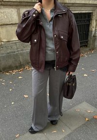 Personne portant une veste en cuir marron, un t-shirt henley gris, un pantalon gris à jambes larges, des chaussures noires, tenant un sac à main tressé marron sur un trottoir.