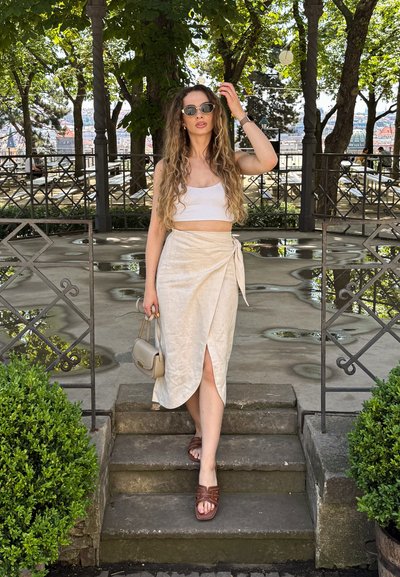Una falda envolvente de un color beige claro con una abertura alta, combinada con un top corto blanco y sandalias marrones. Los accesorios incluyen gafas de sol redondas y un bolso pequeño.
