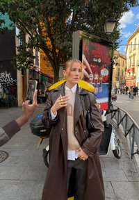 Casaco comprido de couro castanho sobre um suéter claro com um logótipo, por cima de uma camisa branca e calças às riscas. Anéis dourados e mala de ombro preta.