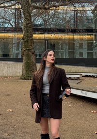 Νεαρή γυναίκα με γκρι πλεκτό ζιβάγκο, μαύρη φούστα και σκούρο παλτό, κρατώντας γυαλιά ηλίου, στέκεται σε εξωτερικό χώρο δίπλα σε δέντρο και σύγχρονο κτίριο.