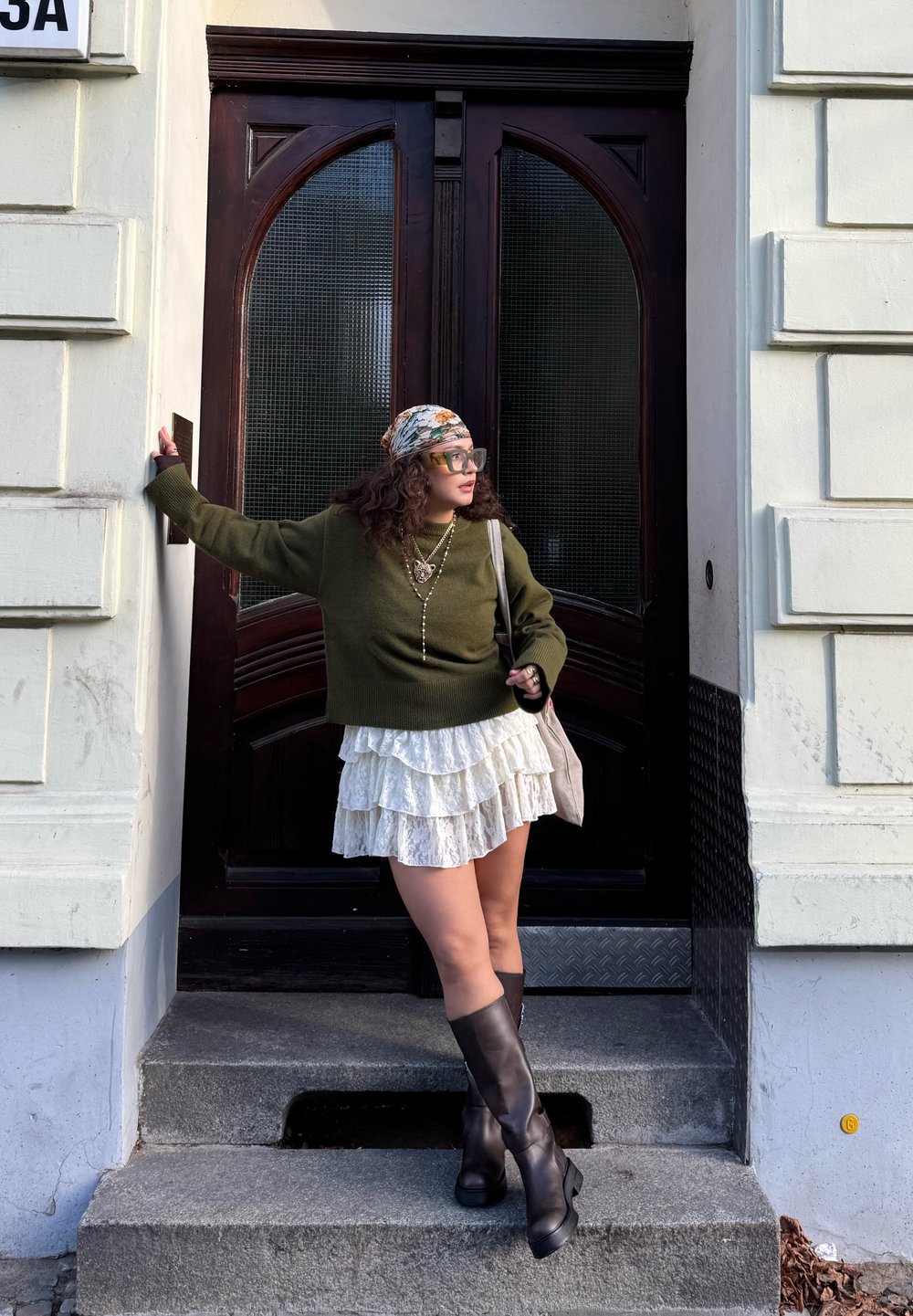 Femme portant un pull vert, une jupe blanche à volants et de hautes bottes noires, debout sur des marches devant des portes doubles en bois sombre.