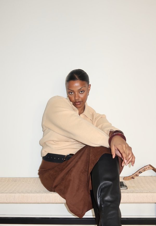 Femme aux cheveux plaqués en arrière, portant un pull beige, une jupe marron et des bottes noires, assise sur un banc tressé contre un mur blanc uni.