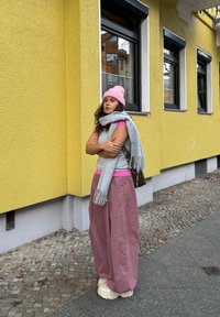 Écharpe gris tricotée, bonnet rose, haut sans manches gris sur une chemise rose, pantalon large rose et baskets blanches épaisses devant un mur jaune.