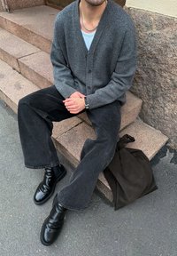 Grey cardigan over white shirt, black wide-leg jeans, and black leather ankle boots. A dark bag is on the ground next to the seated figure.