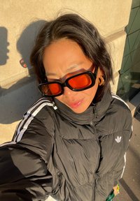 Personne portant une veste noire Adidas à rayures blanches et de grandes lunettes de soleil rectangulaires orange, debout à l'extérieur au soleil.